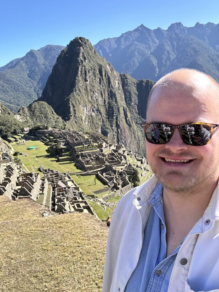 Man glimlacht naar de camera met Machu Picchu op de achtergrond. Het uitzicht toont de ruïnes van de historische Inca-stad, omringd door groene bergen onder een heldere blauwe lucht.